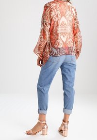 Woman wearing sheer orange patterned blouse, rolled-up light blue jeans, and metallic rose gold ankle-strap block heel sandals, standing facing away.