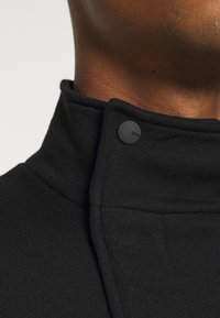 Collet noir en tissu texturé, avec un détail à bouton-pression. Vue en gros plan, mettant en valeur le matériau et les coutures.