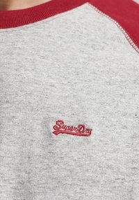 Närbild av grått tyg med röd krage och röd broderad "Superdry"-logotyp på bröstområdet av en skjorta.