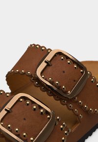 Καφέ δερμάτινα λουριά σανδαλιού με αγκράφες σε χρυσή απόχρωση και διακοσμητικά χρυσά καρφιά κατά μήκος των κυματιστών άκρων.