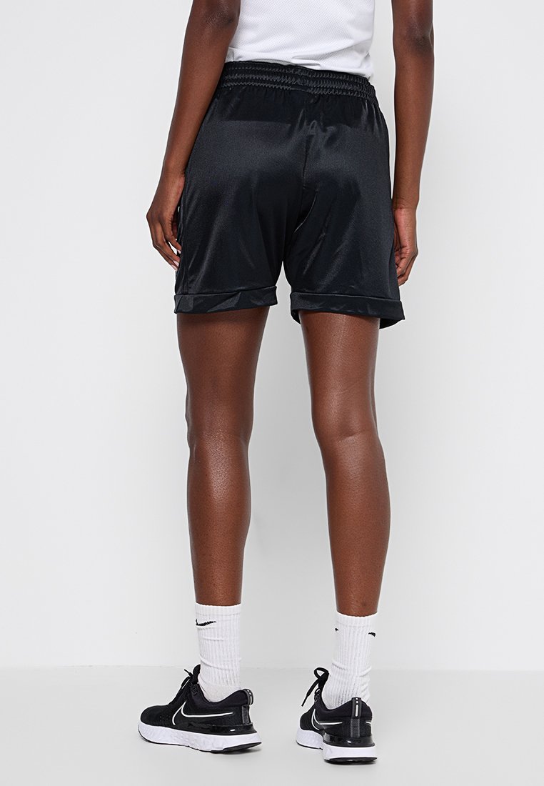 Shorts de sport noirs en satin avec une taille élastique et des fentes sur les côtés, à mi-mollet, accompagnés de chaussettes blanches et de baskets noires.