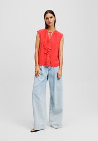 Blusa de color coral con un frente con volantes, diseño sin mangas y escote en forma de llave, combinada con jeans de cintura alta de corte amplio en un lavado claro. Sandalias negras simples.