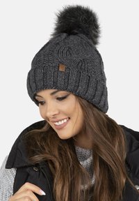 Beanie gri tricotat cu un pom-pom mare din blană pe vârf. Prezintă un model de tricot cu cabluri și o etichetă mică din piele maro pe partea din față.