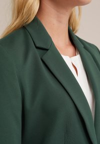 Blazer verde scuro con un design aderente, caratterizzato da un colletto a risvolto, spalle strutturate e tessuto testurizzato. Colletto della camicia bianco visibile sotto.