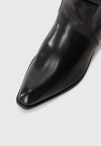 Bottine en cuir noir à bout pointu, texture lisse et détails de couture minimaux. La semelle a une légère élévation.