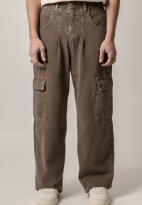 Rudos darbo kelnės su laisvu siluetu, turinčios du šoninius kišenes, matomus siūlus ir klasikinį sagos užsegimą.