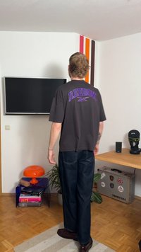 Tamsiai pilka Reebok marškinėlių su violetiniu logotipu, derinta su tamsiai mėlynomis džinsais ir rudais batais. Kambaryje yra televizorius, ryški siena ir dekoracijos.