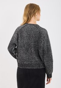 Melnbalts adīts džemperis ar teksturētu, zigzag tipa rakstu, raglan piedurknēm un ribotu apakšmalu, valkāts virs melna svārka.