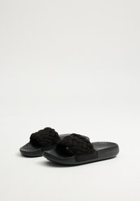Chanclas negras de slide con correas de tejido entrelazado, suelas texturizadas negras y una sensación suave y acolchada. El diseño presenta un estilo minimalista.