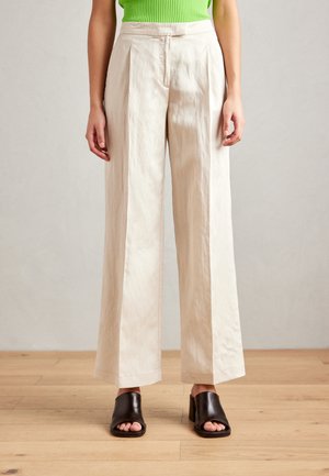 Wide-leg trousers in pale beige, made of soft fabric. Features front pleats, a high waist, and a relaxed fit. Paired with black open-toe mules.