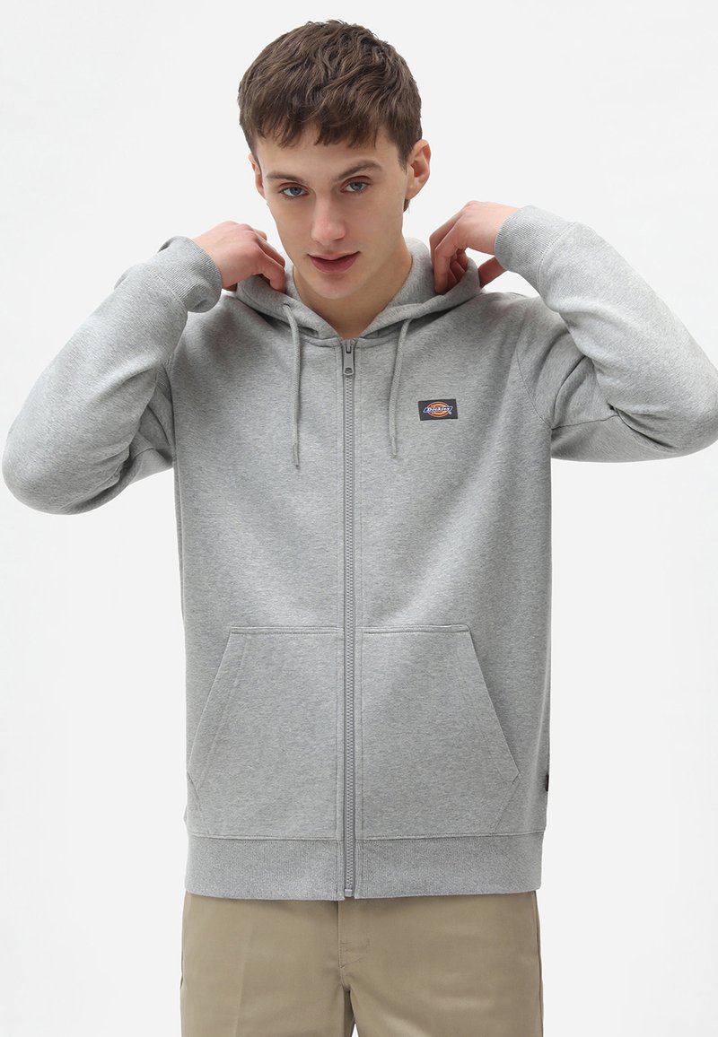Gray zip-up hoodie made of soft fabric with a front pocket, drawstrings, and a small logo patch on the left chest.
