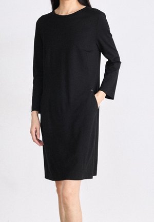 Femme portant une robe noire simple à manches longues, longueur genou, avec un col rond, une main dans la poche, debout devant un fond uni.