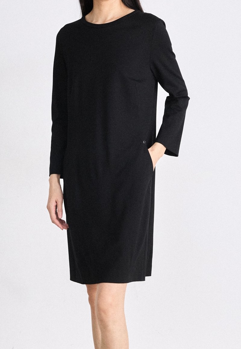 Femme portant une robe noire simple à manches longues, longueur genou, avec un col rond, une main dans la poche, debout devant un fond uni.