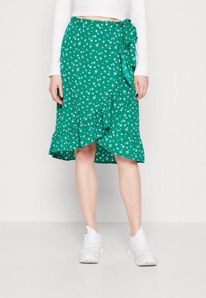 Jupe portefeuille verte avec petit imprimé floral blanc et ourlet à volants, portée avec un haut blanc à manches longues et des chaussures de sport blanches.