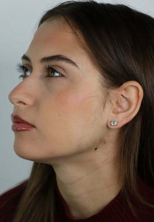 Jonge vrouw met bruin haar en lichte make-up die omhoog kijkt, met een enkele diamanten oorbel in haar linker oor.