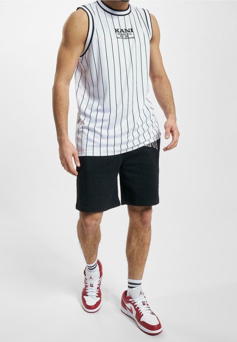 Chemise blanche sans manches avec des rayures verticales noires et un logo, associée à un short noir et des chaussures de sport rouges et blanches.