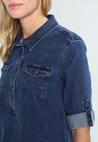 Camisa de denim azul oscuro con cuello abotonado, bolsillos en el pecho y mangas enrolladas. Presenta costuras prominentes y una textura suave.
