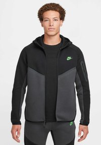 Sudadera con cremallera de Nike en negro y gris con un logo verde. Presenta un diseño ajustado, bolsillo con cremallera en la manga y una textura suave.