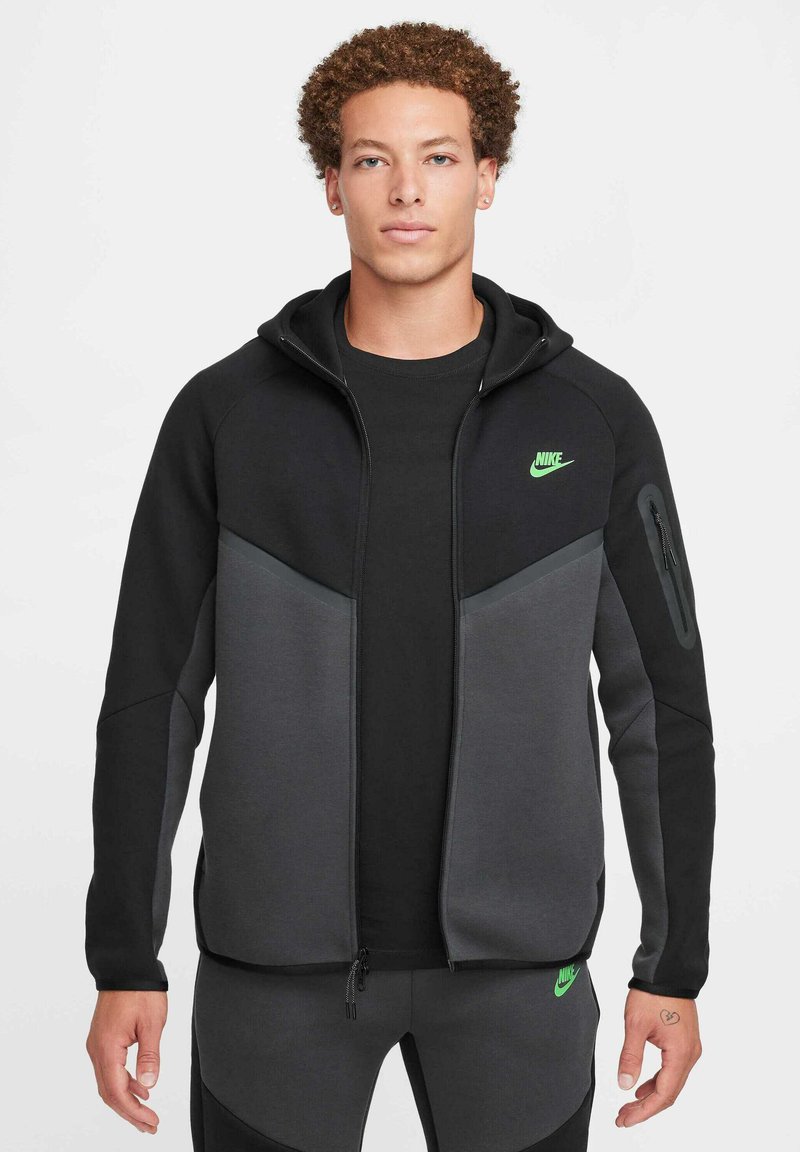 Sudadera con cremallera de Nike en negro y gris con un logo verde. Presenta un diseño ajustado, bolsillo con cremallera en la manga y una textura suave.