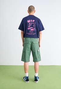 Navy blue t-shirt with pink graphic on back, paired with green cargo shorts featuring multiple pockets. Model wears sneakers with white socks.