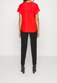 Kobieta w luźnej czerwonej bluzce z krótkim rękawem, dopasowanych czarnych spodniach i czarnych botkach na obcasie, stojąca na jednolitym jasnym tle.