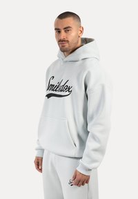 Felpa con cappuccio grigia chiara realizzata in un tessuto morbido e texture. Presenta una tasca frontale, un logo ricamato nero e un cappuccio regolabile con cordino. Look elegante e vestibilità rilassata.