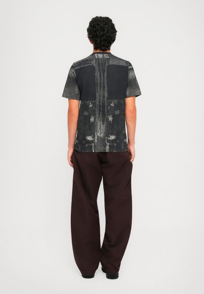 Man wearing black and gray abstract-print T-shirt and loose dark brown pants, standing facing a plain white wall.