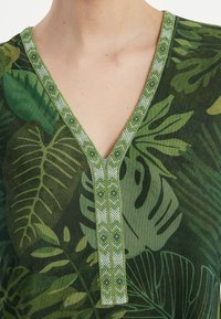 Grünes V-Ausschnitt-Top mit einem Blättermuster, strukturiertem Stoff und verziertem Saum mit geometrischen und floralen Perlendetails in passenden Farben.