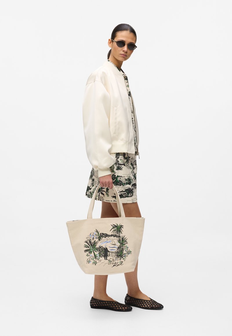 Γυναίκα που φοράει κρεμ μπουφάν μπότερμα και φλοράλ φόρεμα, κρατώντας μπεζ τσάντα ώμου με εικονογράφηση τροπικού τοπίου και μαύρες πλεκτές εσπαντρίγιες.