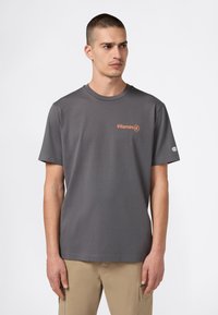 Mężczyzna z krótkimi włosami, ubrany w szary t-shirt z pomarańczowym napisem "Witamina C" oraz beżowe spodnie cargo, stojący na tle białym.