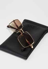 Lunettes de soleil avec des verres carrés marron, une monture en métal doré et des embouts de branches noirs, reposant sur une pochette noire avec "QUAY" en relief.