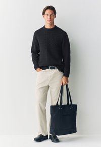 Čierny pletený sveter s textúrovanými vzormi, béžové nohavice s rovnými nohavicami a čierna kabelka s rúčkami. Modelka stojí s rukami v vreckách, obutá v čiernych topánkach.