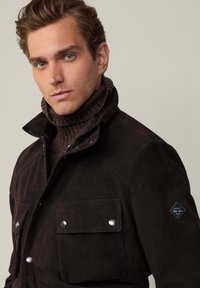 Donkerbruin suede jack met een kraag, zilveren drukknoopjes, twee voorzakken en een blauwe logo patch op de schouder. Geribbelde coltrui eronder.
