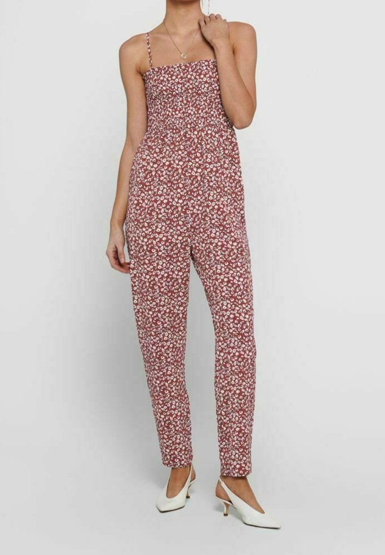 Femme portant une combinaison sans manches à fleurs couleur rouille avec fines bretelles et des chaussures slingback blanches à petit talon, debout devant un fond uni.