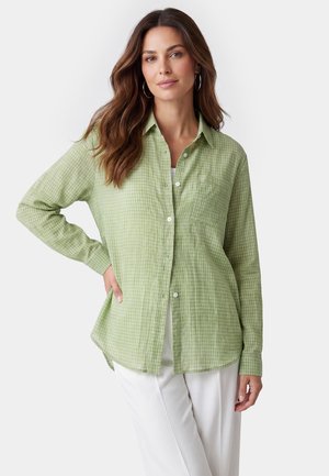Femme aux longs cheveux bruns portant des boucles d’oreilles argentées en forme d’anneau, une chemise à carreaux vert clair boutonnée et un pantalon blanc sur un fond uni.