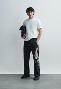 Λευκό κοντομάνικο μπλουζάκι με κόκκινη κεντημένη επιγραφή. Μαύρο παντελόνι φόρμας με πολύχρωμα αφηρημένα σχέδια. Το μοντέλο κρατάει μια μαύρη τσάντα.