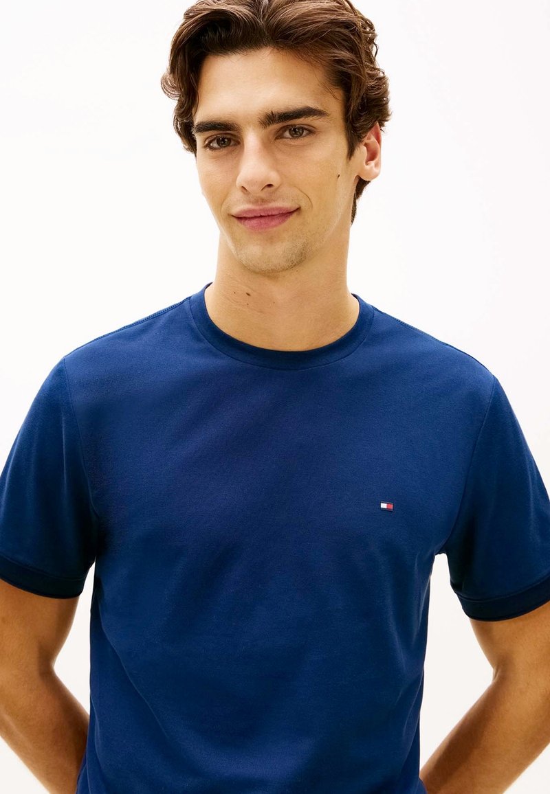 Jeune homme aux cheveux bruns portant un t-shirt bleu marine uni avec un petit logo Tommy Hilfiger sur la poitrine, souriant légèrement.