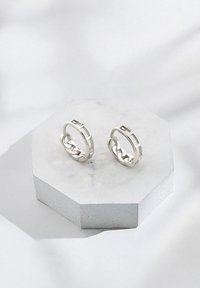 Silverfärgade ringörhängen med kedjelänksdesign, med släta ytor och geometriska detaljer, visade på en sexkantig marmorställ.