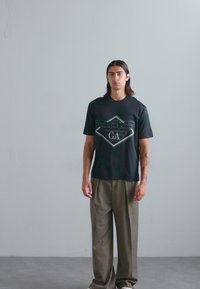 Zwarte t-shirt met een geometrisch grafisch ontwerp en centraal logo, gecombineerd met losse beige broek. Eenvoudige ronde halslijn, korte mouwen.