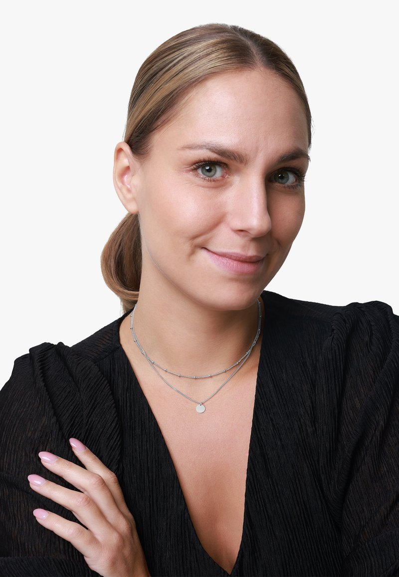 Silberne, geschichtete Halsketten mit einem kleinen Anhänger und einer strukturierten Kette, getragen von einem Modell mit glattem, hellbraunem Haar und einem schwarzen Oberteil.
