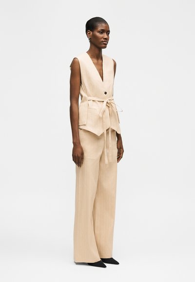 Femme portant un gilet beige sans manches avec une ceinture à nouer à la taille et un pantalon large assorti, debout devant un fond clair uni.