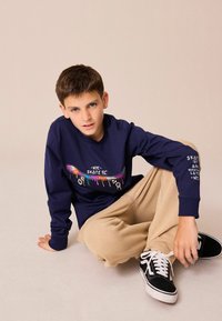 Tengerészkék hosszú ujjú póló élénk színekben skateboard grafikával, bézs bő nadrággal és fekete sportcipővel, fehér fűzőkkel.