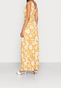 Robe maxi jaune moutarde avec des motifs floraux blancs, sans manches, et dans le dos en V. Portée avec des sandales à talons blanches.
