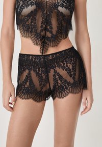 Conjunto de bralette y pantalones cortos de encaje negro con patrones florales, bordes ondulados y textura transparente, diseñado para una silueta ajustada.