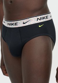 Κοντινή λήψη ενός ατόμου που φοράει μαύρα εσώρουχα Nike με πολύχρωμη λευκή ζώνη μέσης με κηλίδες, που φέρει λογότυπα Nike και κίτρινο λογότυπο swoosh στο μπροστινό μέρος.