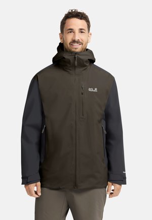 Man die een bruin-zwarte Jack Wolfskin jas met ritssluiting en capuchon draagt, staand en glimlachend tegen een effen achtergrond.