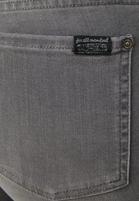 Un bolsillo trasero de mezclilla gris con una etiqueta de tela negra cosida en la parte superior y un remache metálico en la esquina superior derecha.