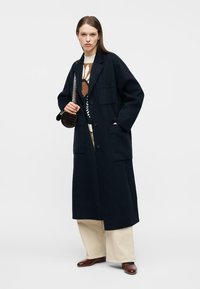 Woman wearing a long black coat, patterned sweater, beige wide-leg pants, brown shoes, and holding a small dark handbag on shoulder.