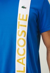 Modra polo majica iz teksturirane tkanine, s širokim belim pasom na katerem je v rumeni barvi napis "LACOSTE" ter majhnim zelenim krokodilom.