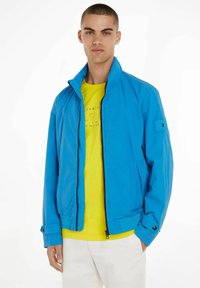 Leichte blaue Jacke mit Stehkragen, Reißverschluss und geknöpften Manschetten, getragen über einem leuchtend gelben Hemd. Glatte Textur, lässiges Design.